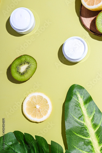 Natural skincare cream jars placed with fresh lemon and kiwi slices, large tropical leaves, and wooden props on a pastel lime background. Bright, healthy, organic beauty product concept