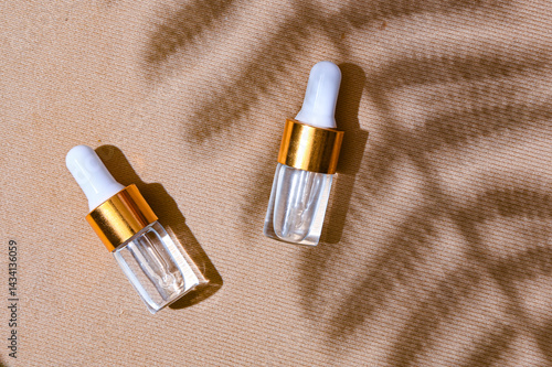 Two transparent dropper serum bottles with white caps and gold collars placed on a textured beige background with soft leaf shadows. Natural skincare branding concept with summer mood