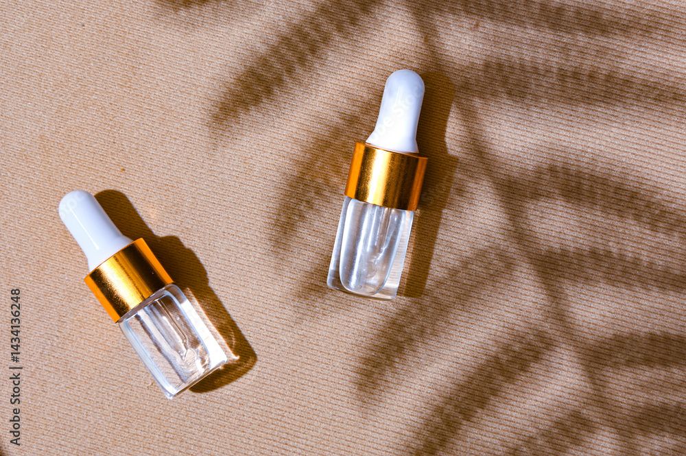 Two transparent dropper serum bottles with white caps and gold collars placed on a textured beige background with soft leaf shadows. Natural skincare branding concept with summer mood