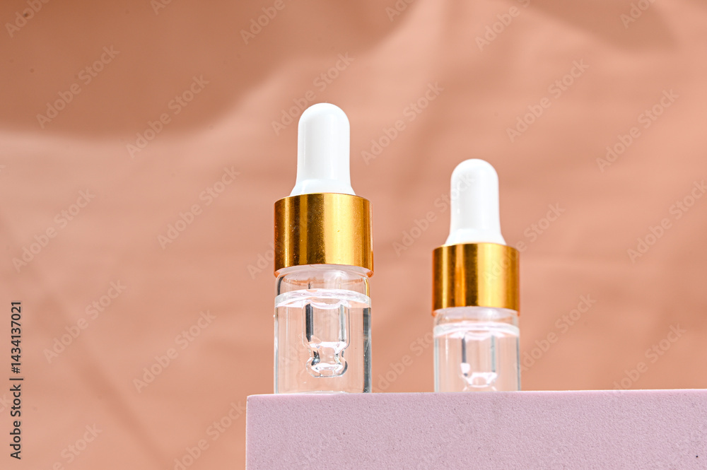 Two transparent glass serum dropper bottles with white tips and golden collars placed on a soft pink pedestal, against a peach background. Minimalist skincare branding with clean aesthetics