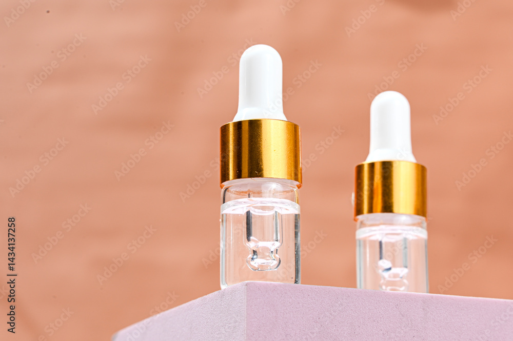 Two transparent glass serum dropper bottles with white tips and golden collars placed on a soft pink pedestal, against a peach background. Minimalist skincare branding with clean aesthetics