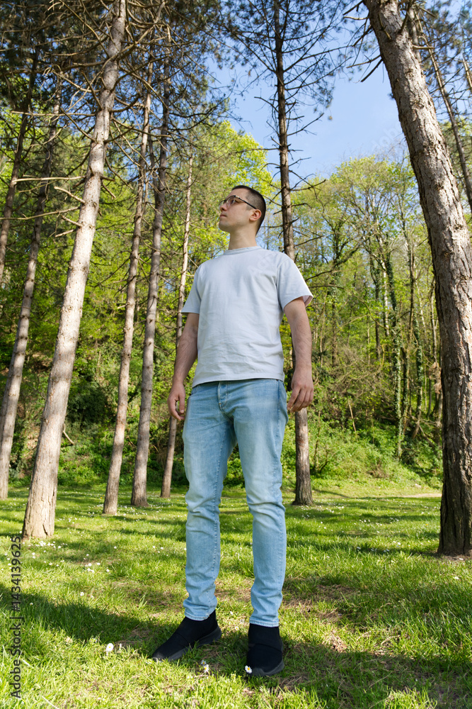 A man stands in a serene forest clearing, surrounded by lush greenery and tall trees, wearing a white t-shirt, light blue jeans, and black shoes, gazing upwards A man stands in a serene forest clearing, surrounded by lush greenery and tall trees, wearing a white t-shirt, light blue jeans, and black shoes, gazing upwards