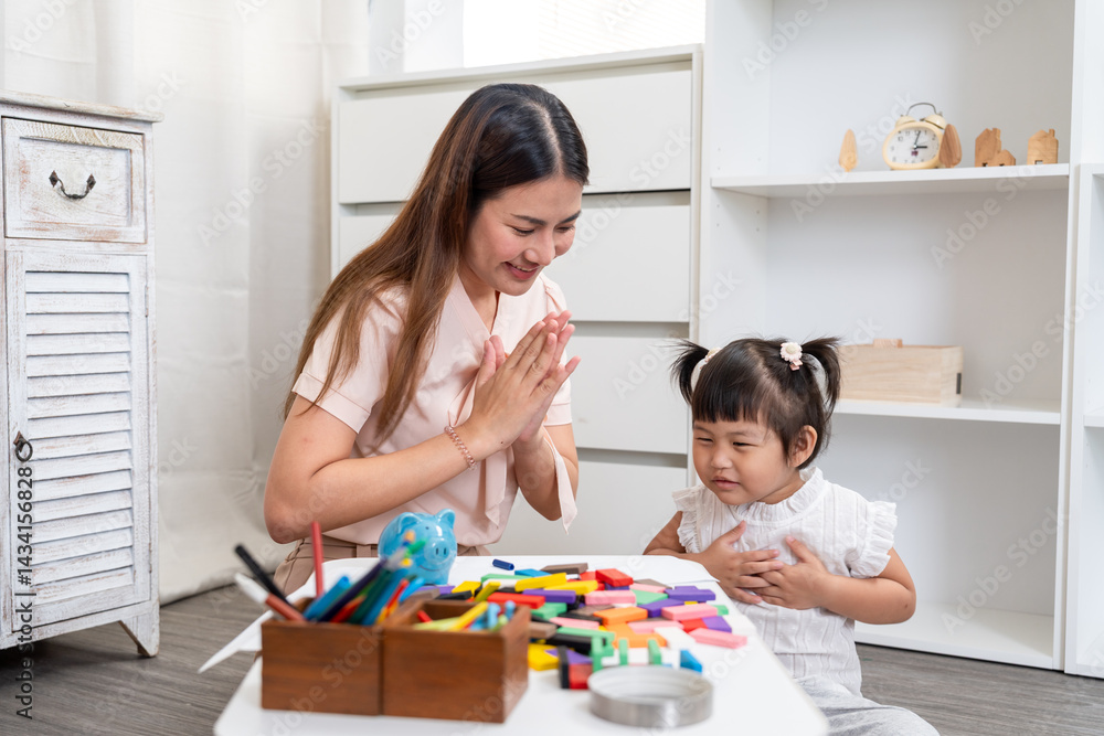 Mother and preschool daughter sitting on floor practicing gratitude gesture hands together during educational activity at home play area with colorful blocks and blue piggy bank on white table. Mother and preschool daughter sitting on floor practicing gratitude gesture hands together during educational activity at home play area with colorful blocks and blue piggy bank on white table.