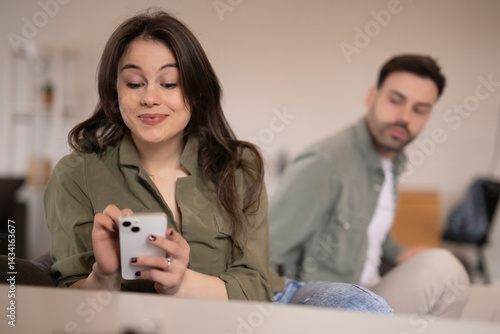 Young woman smiling at smartphone while man in background looks concerned

