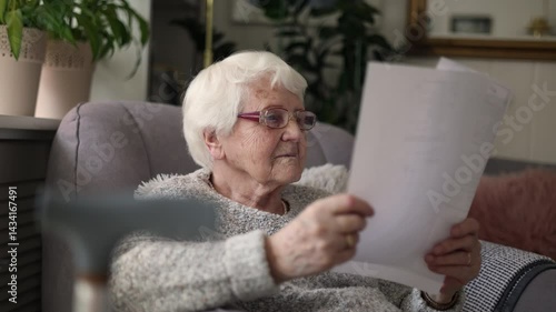 Elderly woman wearing eyeglasses reading documents while sitting in an armchair in living room