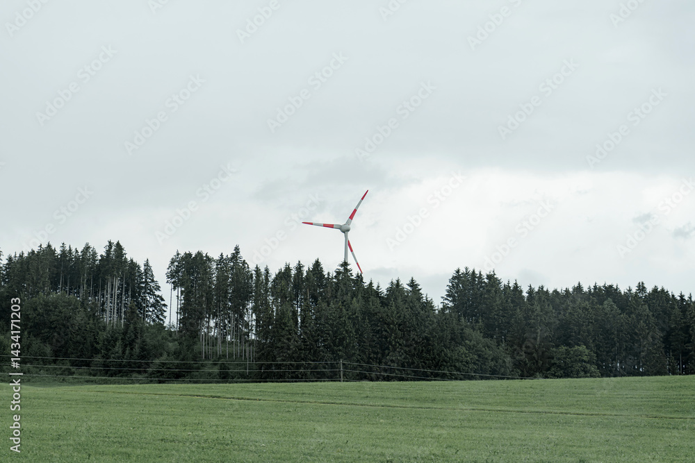 Fototapeta premium Wind turbine spins gracefully in a cloudy landscape surrounded by lush green trees