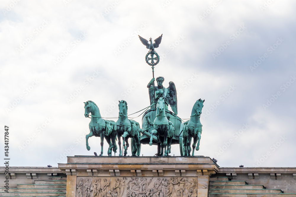 Obraz premium The Brandenburg Gate in Berlin features a majestic statue of a chariot led by four horses, symbolizing peace and victory against a cloudy sky backdrop.