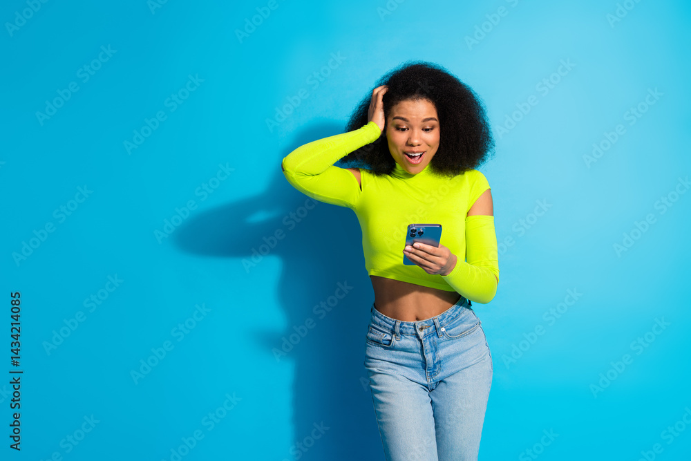 Young woman in stylish neon green top using smartphone against blue background expressing joyful emotions with casual pose