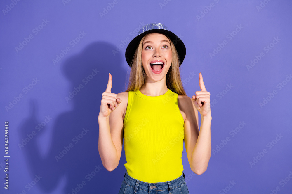 Young woman in a cheerful pose wearing stylish clothes and hat, gesturing upward with a joyful expression on a vibrant purple background