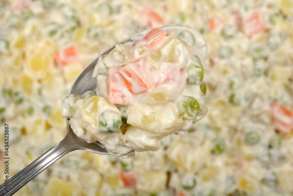 A spoonful of creamy Croatian-style French salad with diced vegetables and mayonnaise is held over the remaining salad in a blurred background