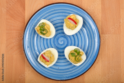 Obraz na płótnie A top-down view shows four deviled eggs, filled with a creamy avocado and yolk mixture and garnished with green onions and radish slices, arranged on a blue swirled plate on a wooden surface w salonie