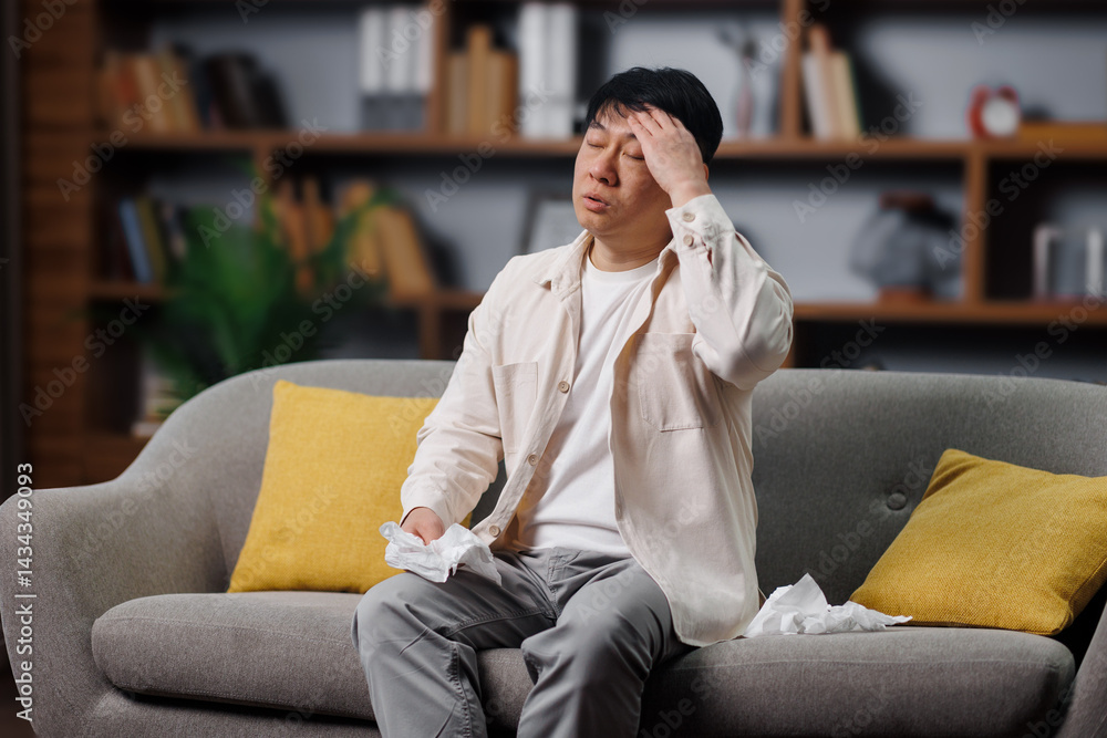 ill sick man, feeling unwell, having headache, man is seated on a couch in a cozy living room, visibly distressed while wiping his forehead with a tissue. Several decorative bookshelves and greenery ill sick man, feeling unwell, having headache, man is seated on a couch in a cozy living room, visibly distressed while wiping his forehead with a tissue. Several decorative bookshelves and greenery