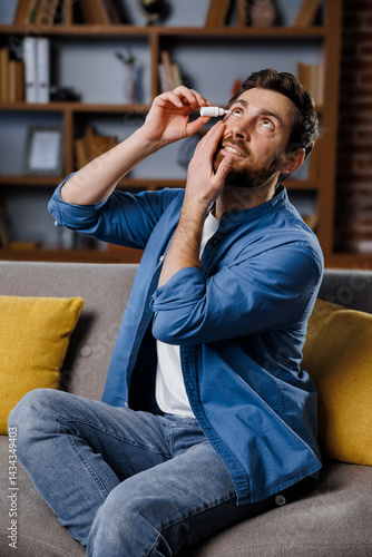 A man is seated comfortably on a sofa in a cozy living room, carefully applying eye drops to his eye. Soft lighting enhances the relaxed atmosphere, indicating an evening routine.