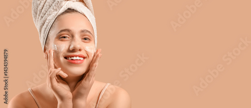 Young woman in towel applying facial cream on beige background, closeup