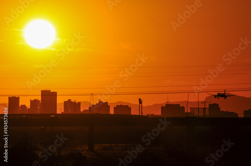 A commercial airplane approaches Sky Harbor International Airport as the sun sets behind the Phoenix skyline, viewed from Tempe Town Lake, illustrating the intense heat of Arizona's Valley of the Sun