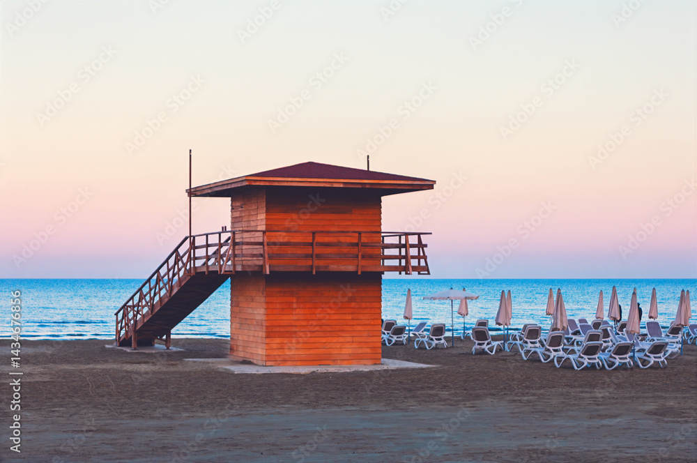 Wooden lifeguard tower stands on tranquil beach surrounded by lounge chairs and closed umbrellas. With pastel sky and calm sea as its backdrop, serene scene evokes early morning or evening tranquility