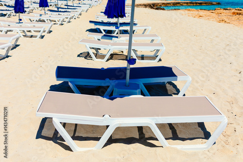 Obraz na płótnie Rows of white beach chairs are neatly arranged on a sandy beach under blue umbrellas, casting shadows in sunlight. In background turquoise sea meets rocky cliffs, creating serene coastal scene w salonie