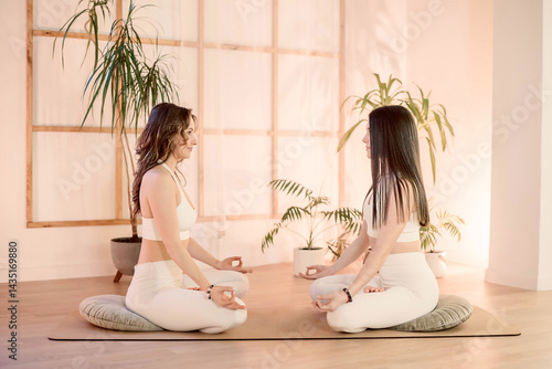 Two women sit cross-legged facing each other in soft light, connection through synchronized breath and mutual focus, connection deepens in tranquil yoga studio atmosphere bond captured composition 