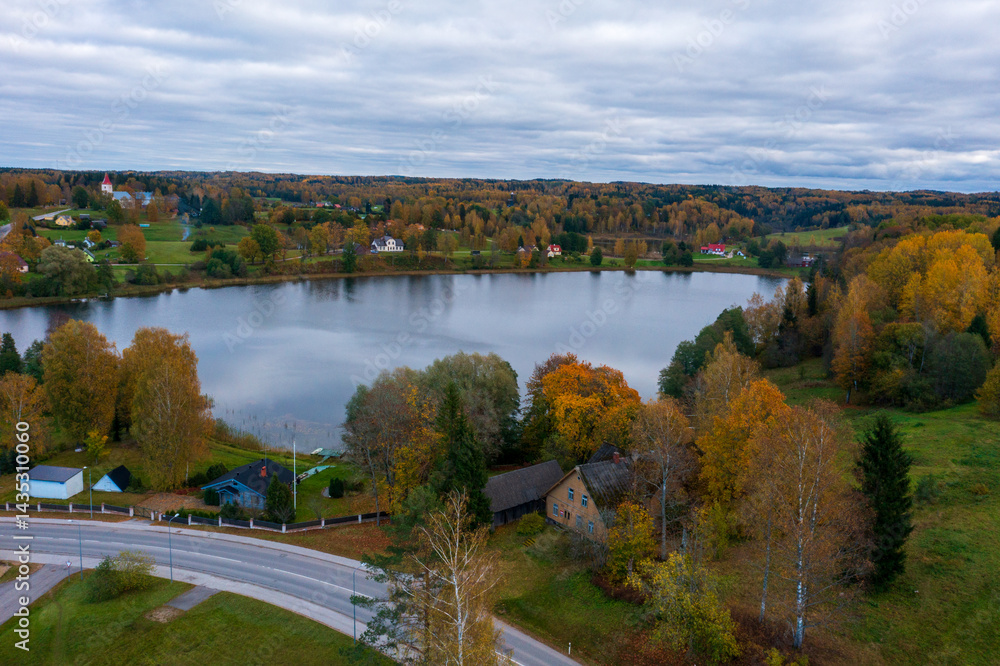 Fototapeta premium Aerial view of Rõuge lake surrounded by vibrant autumn foliage in the Estonian countryside. Rural village with scattered houses along the shoreline under cloudy skies revealing the beauty of Estonia