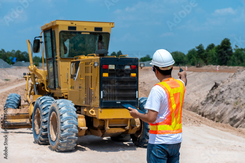 фотографія Construction Worker Operating Grader: A construction worker in a safety vest and helmet operates a large yellow grader, skillfully maneuvering the machine on a construction site