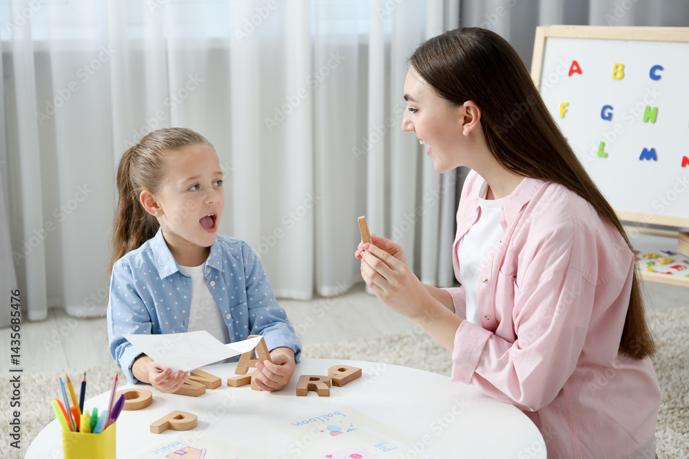 Fototapeta premium Speech therapist teaching little girl alphabet with wooden letters at white table indoors