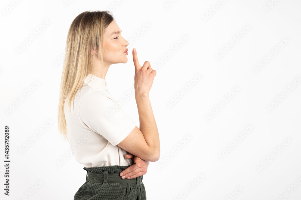 Woman standing profile the camera and showing silence gesture with finger. Model wearing white blouse and khaki pants. White studio background Woman standing profile the camera and showing silence gesture with finger. Model wearing white blouse and khaki pants. White studio background