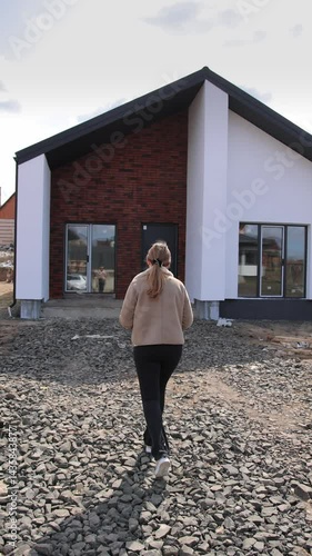Vertical video. A woman walks towards a newly constructed home surrounded by gravel and landscaping. The modern architecture stands out against a clear sky, showcasing new beginnings. Modern duplex.