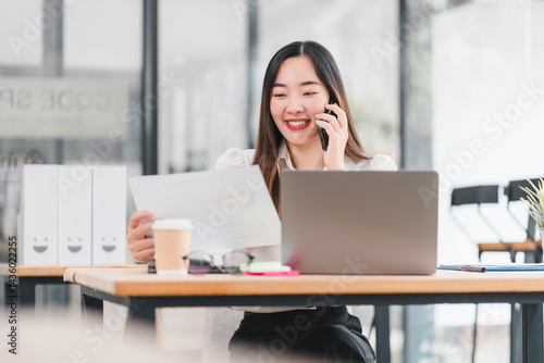 Young woman smiling while talking on phone and working with document and laptop in modern office