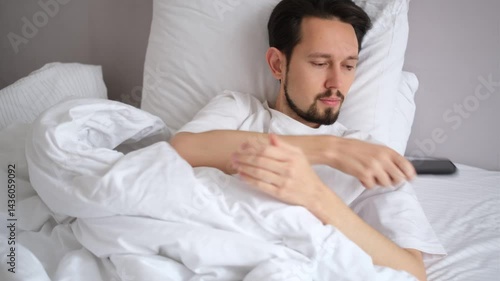 young brunette with a beard just woke up and lying in bed watching the news on his phone