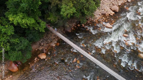 Rustic wooden cable bridge spans a tranquil river nestled in green mountains. Lush nature surrounds this peaceful crossing