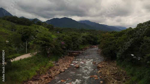 Rustic wooden cable bridge spans a tranquil river nestled in green mountains. Lush nature surrounds this peaceful crossing
