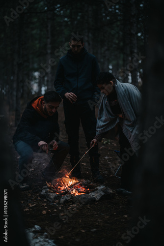 Three friends gathered around a warm campfire in the woods, enjoying a winter picnic atmosphere. They are cooking food and sharing the calmness of a winter evening outdoors.