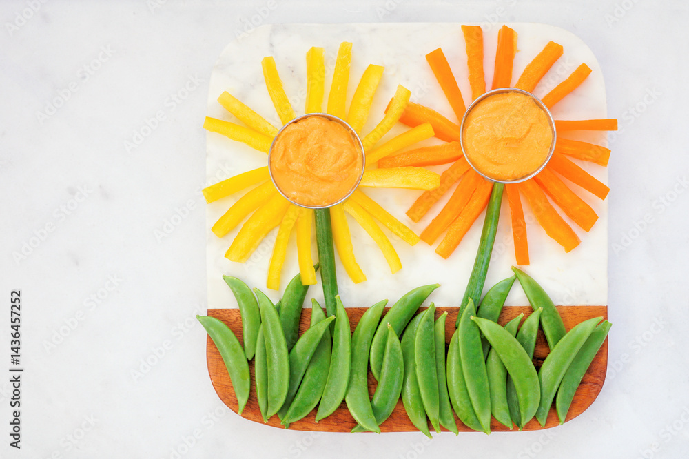 Fun summer theme vegetable snack board against a white marble background. Flower shaped veggies with hummus. Top down view.
