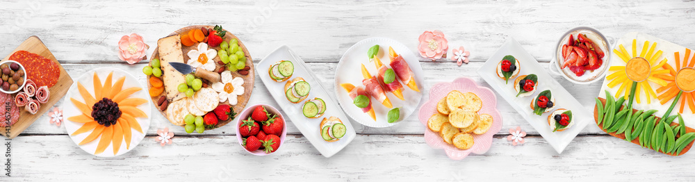 Summer garden party food table scene. Assortment of fun flower theme charcuterie items, cheese, fruit, vegetables and finger snacks. Above view on a white wood banner background.