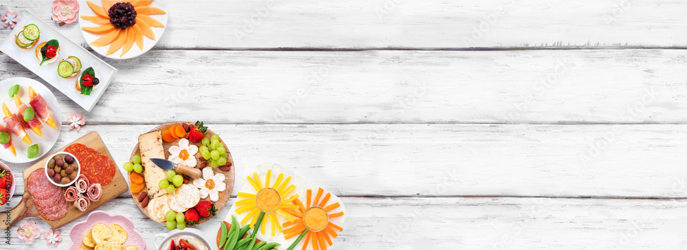 Summer garden party food corner border. Assortment of fun flower theme charcuterie items, cheese, fruit, vegetables and finger snacks. Above view on a white wood banner background.