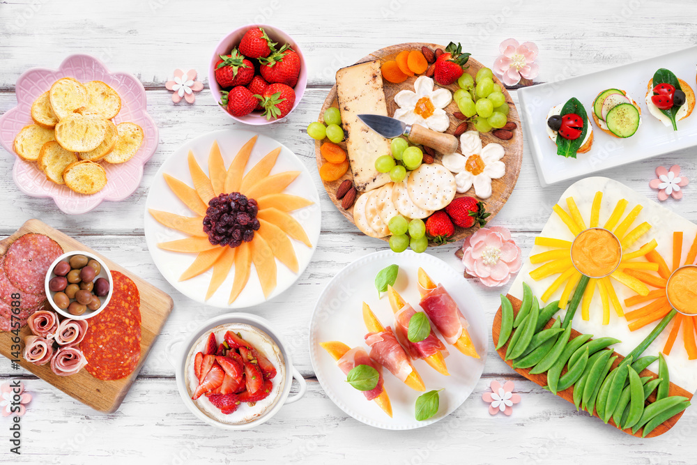 Summer garden party food table scene. Assortment of fun flower theme charcuterie items, cheese, fruit, vegetables and finger snacks. Top down view on a white wood background.