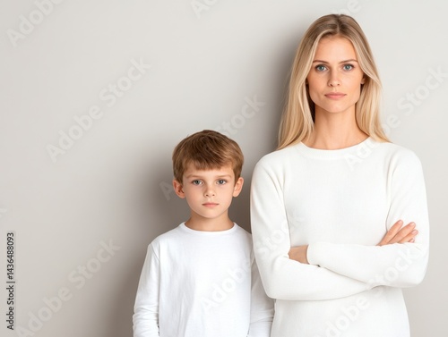 A woman and young boy stand against a neutral background, both wearing white long-sleeved shirts.  They have serious expressions