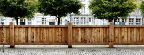 The sturdy wooden fence stands prominently, dividing a peaceful cobblestone street from lush green trees, showcasing a tranquil urban setting under clear skies.