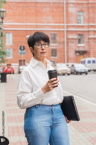 Mature woman with coffee and laptop standing outdoors. Freelance work, career focus and independent lifestyle