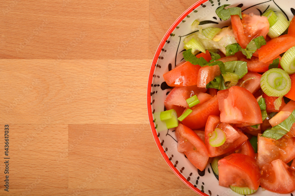 A partially visible colorful bowl of fresh tomato salad with green onions and basil sits on a wooden surface