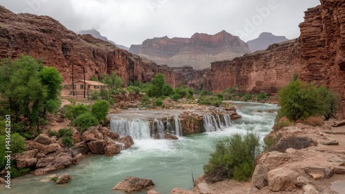 Scenic waterfall cascading in desert canyon with turquoise river and red rock formations on a cloudy day, small house beside the river.