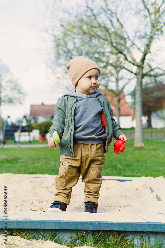 A boy of 2 years old in a spring jacket and hat plays with sand in a sandbox. A boy plays on a playground with a shovel in the sand. A child's walk in the fresh air