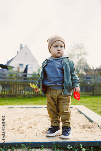 Obraz na płótnie A boy of 2 years old in a spring jacket and hat plays with sand in a sandbox. A boy plays on a playground with a shovel in the sand. A child's walk in the fresh air w salonie