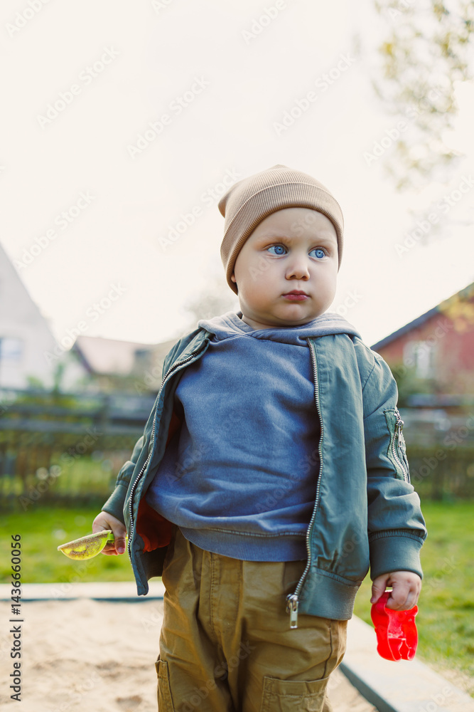 A boy of 2 years old in a spring jacket and hat plays with sand in a sandbox. A boy plays on a playground with a shovel in the sand. A child's walk in the fresh air