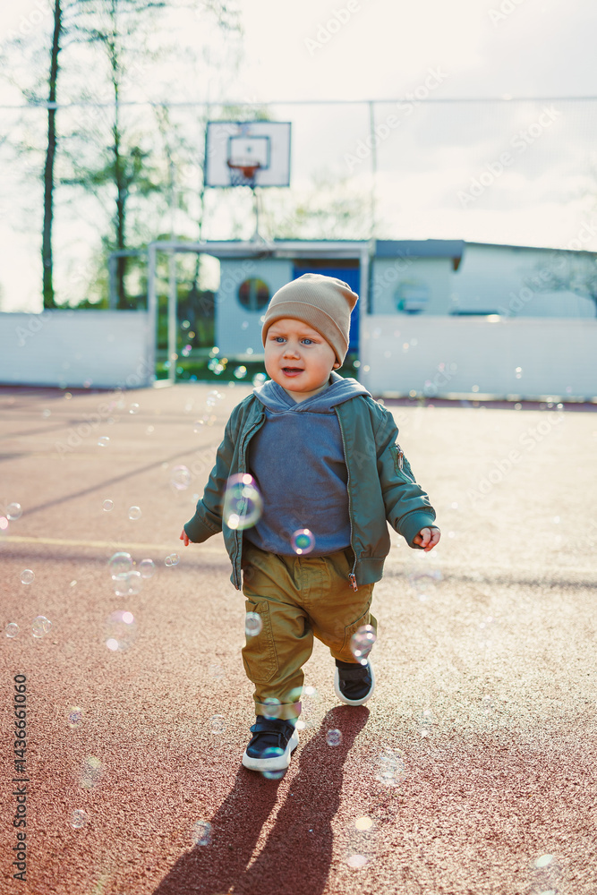 Toddler boy running outside with soap bubbles. Child on a warm spring day for a walk in a jacket and hat.
