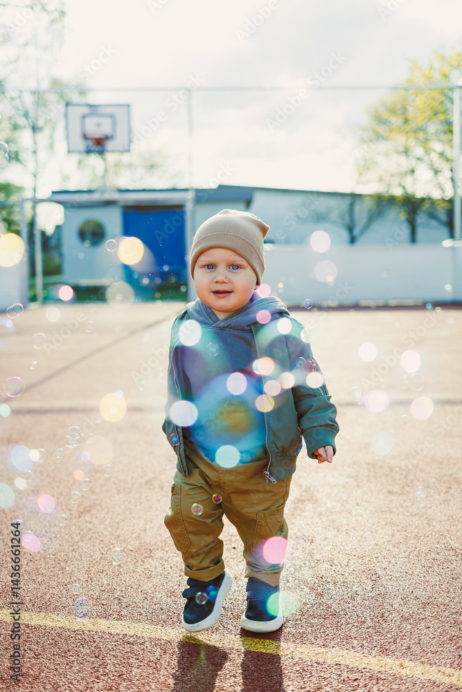 Toddler boy running outside with soap bubbles. Child on a warm spring day for a walk in a jacket and hat.