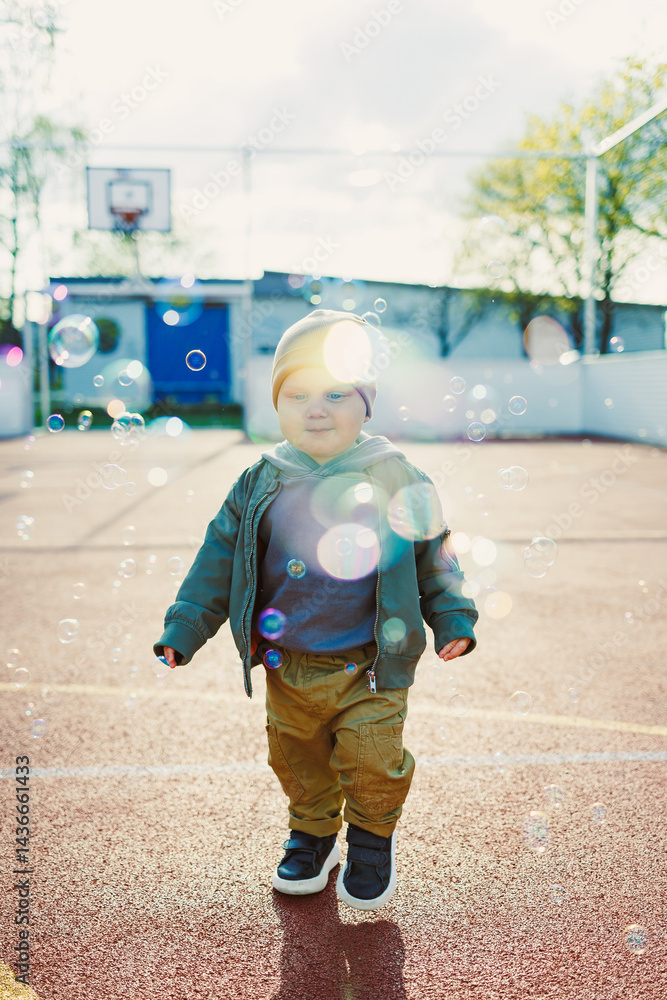 Toddler boy running outside with soap bubbles. Child on a warm spring day for a walk in a jacket and hat.
