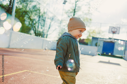 Obraz na płótnie Cheerful 2 year old boy playing outside in summer in a green jacket with soap bubbles. Toddler boy running outside and playing with bubbles. w salonie