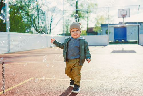 Obraz na płótnie Cheerful 2 year old boy playing outside in summer in a green jacket with soap bubbles. Toddler boy running outside and playing with bubbles. w salonie