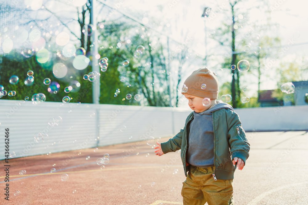 Cheerful 2 year old boy playing outside in summer in a green jacket with soap bubbles. Toddler boy running outside and playing with bubbles.